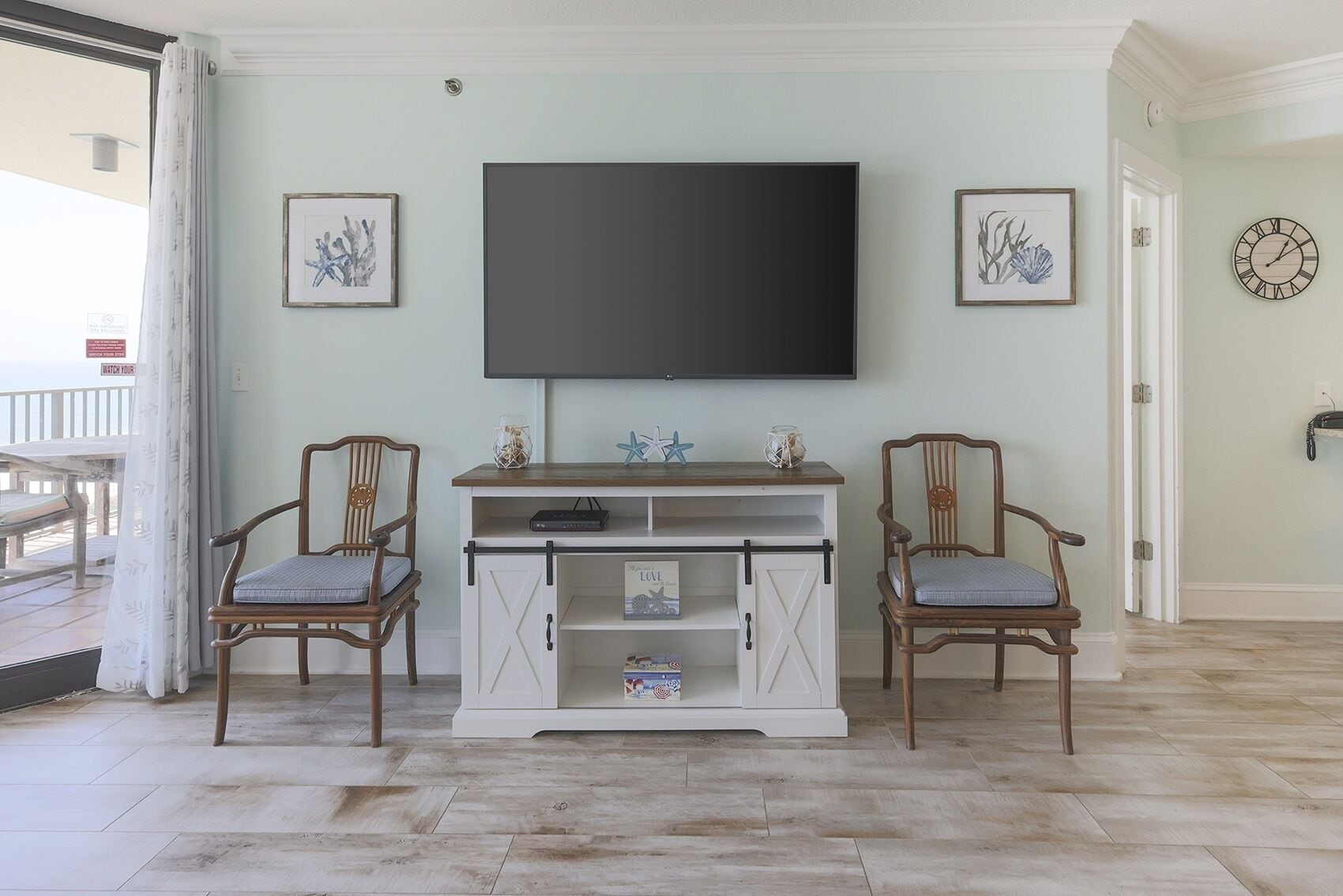 A living room with a flat-screen TV mounted on the wall, two wooden chairs, a white entertainment unit with a shelf, and framed artwork on the walls.