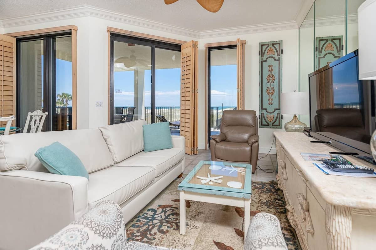 A living room with a sofa, a glass coffee table, a TV, and a view of the beach.