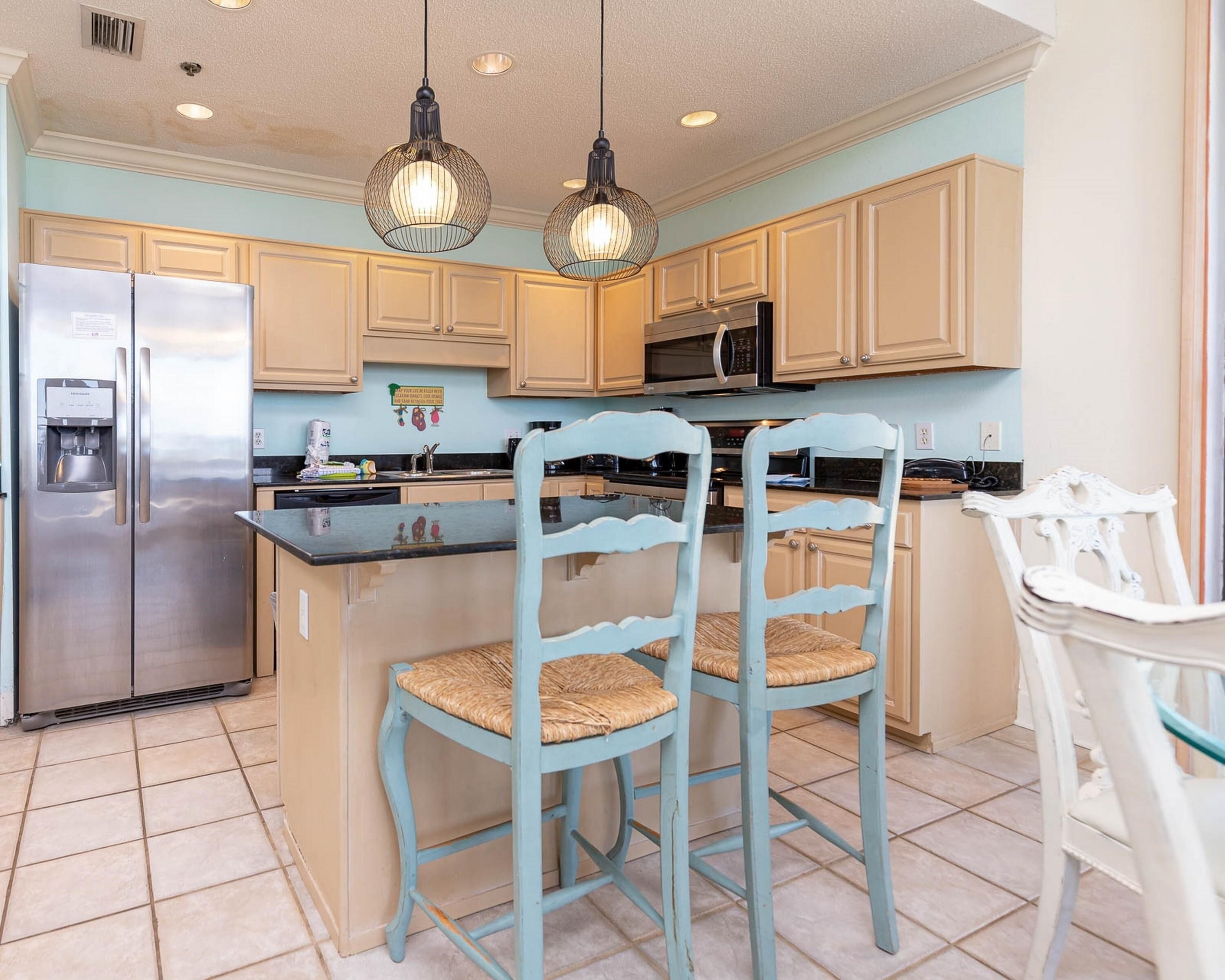 A kitchen with a stainless steel refrigerator, a microwave, and a coffee maker. There are two light blue chairs with woven seats and a white chair with a glass top.