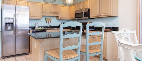 A kitchen with a stainless steel refrigerator, a microwave, and a coffee maker. There are two light blue chairs with woven seats and a white chair with a glass top.