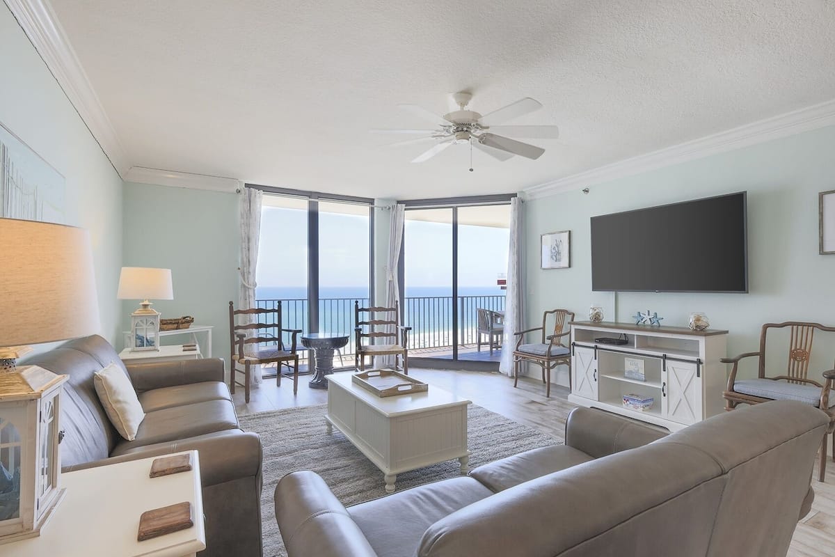A spacious living room with a large flat-screen TV, comfortable seating, and a balcony with a view of the ocean.