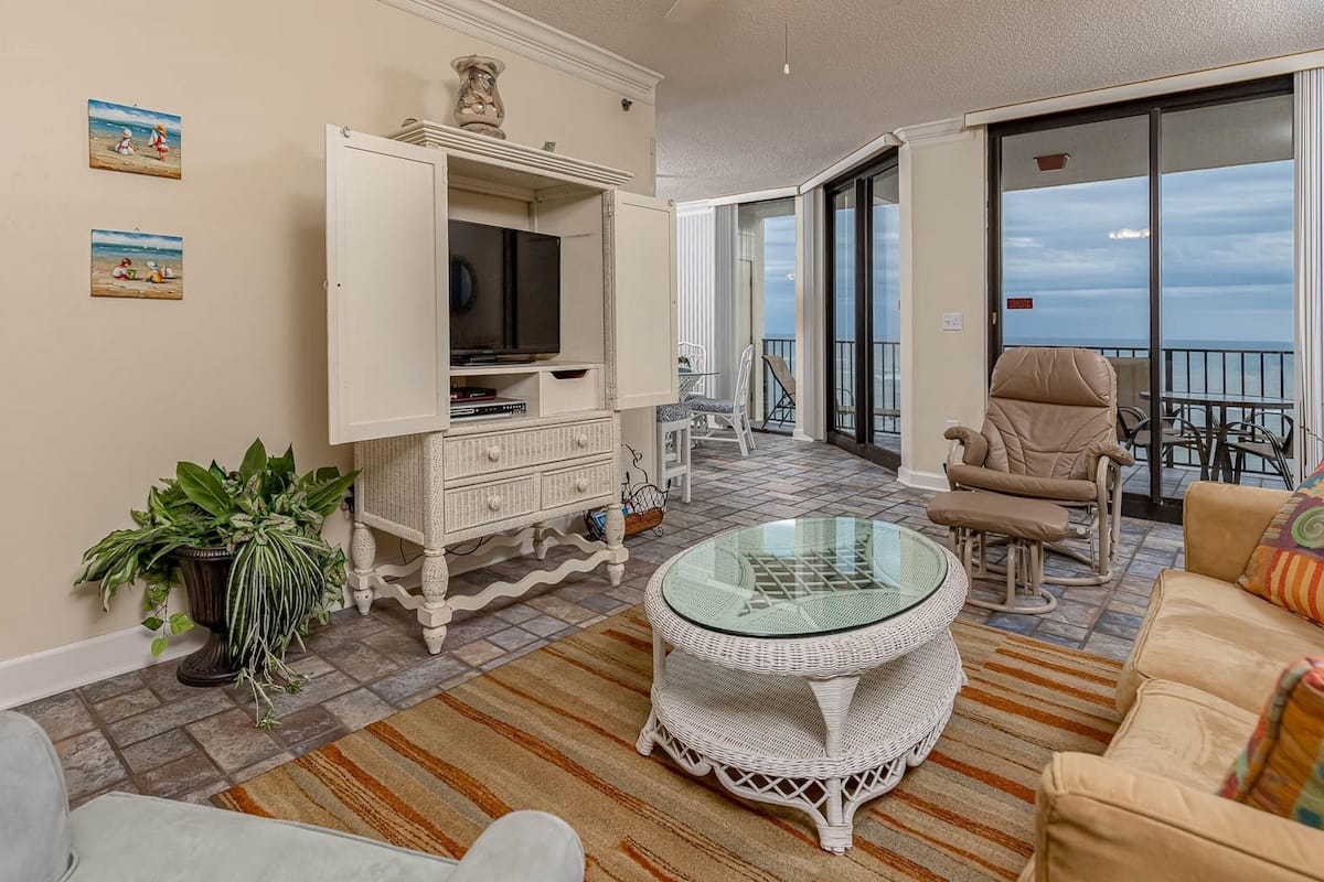 A living room with a television, wicker furniture, a glass coffee table, and a view of the ocean.