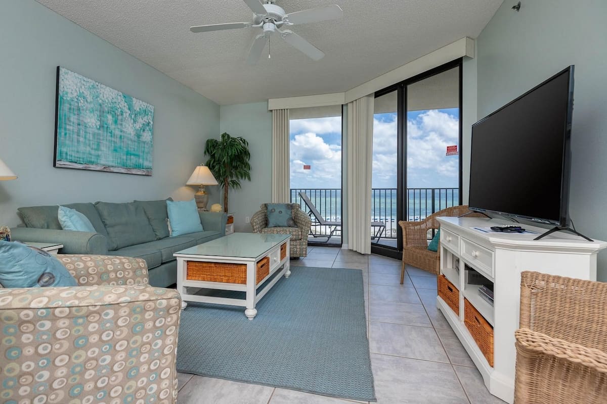 A living room with a sofa, armchairs, a coffee table, a TV, and a balcony with a view of the ocean.