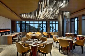Un'area pranzo moderna con diversi tavoli e sedie, ampie finestre che offrono vista all'esterno e un'originale lampada a sospensione.