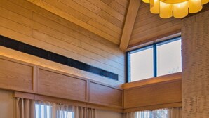 Amplia habitación con techo y paredes de madera, ventanas grandes con cortinas translúcidas, una silla tejida, un sillón de tierno color verde y una mesa redonda de madera.