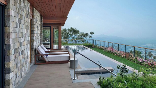 Un espacio exterior moderno con muro de piedra, terraza de madera, sillones de descanso y piscina con vistas panorámicas.
