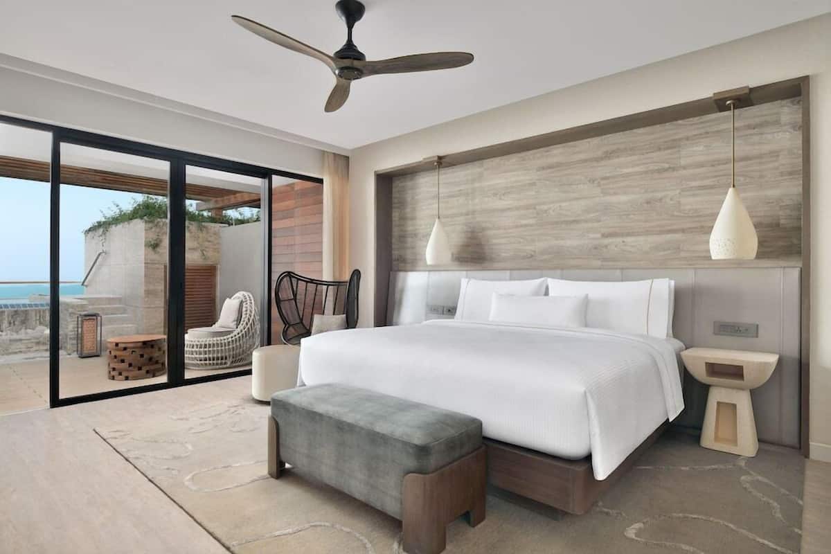 A modern bedroom with a large bed, a ceiling fan, a view of the ocean, and a balcony with outdoor seating.