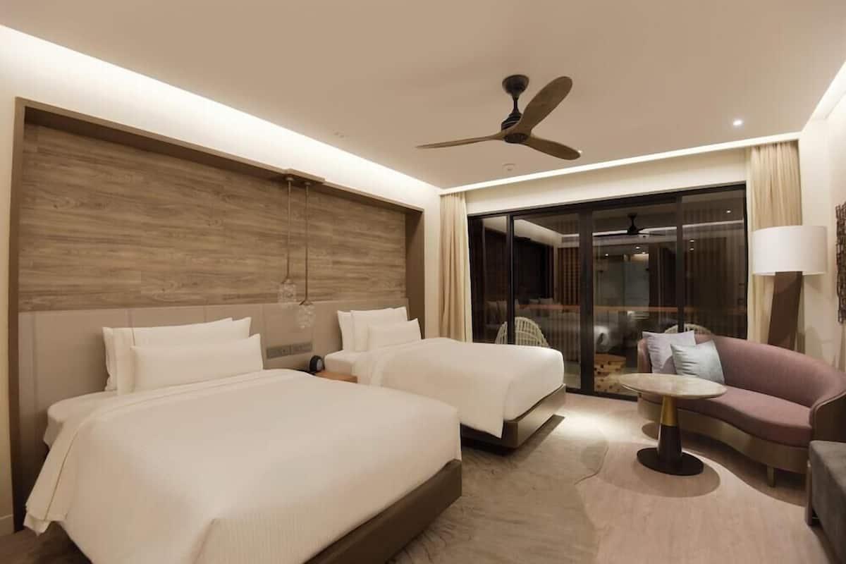 A hotel room with two beds, a sofa, a small table, a ceiling fan, and a sliding glass door leading to a balcony.