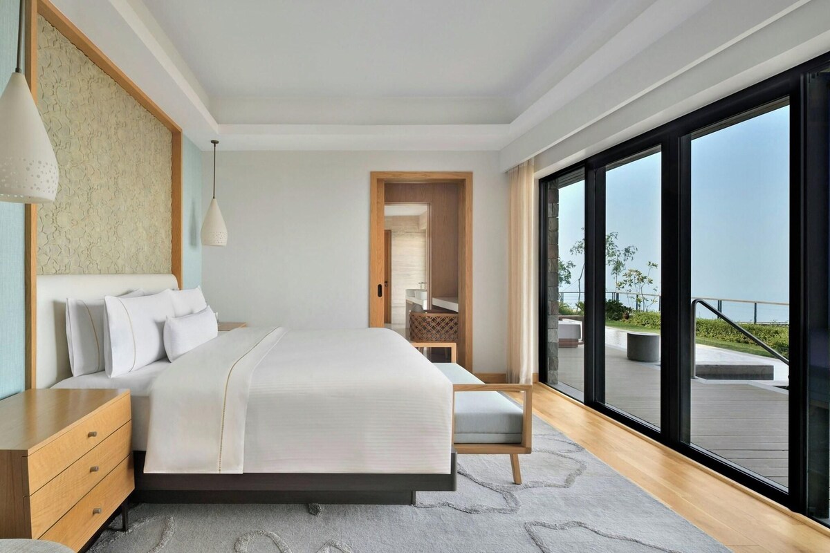 Une chambre à coucher moderne, dotée d’un grand lit, d’une table de chevet, d’une commode et offrant une vue sur l’extérieur grâce à de grandes fenêtres.