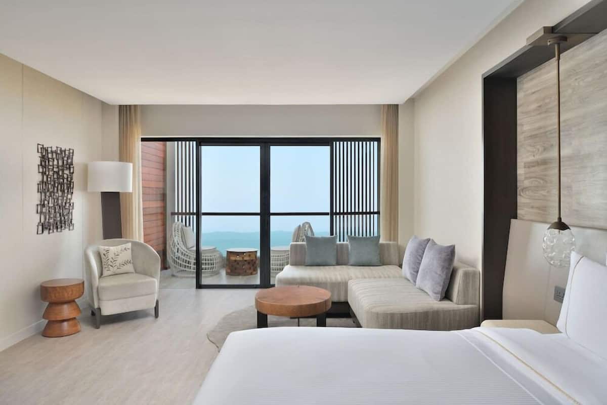 A modern hotel room with a large bed, a sofa, a chair, a round wooden table, and a view through a glass door.