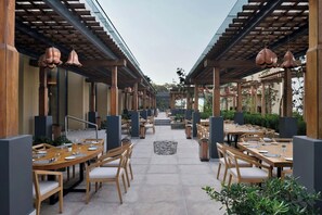 Un espace de restauration en plein air avec des tables et des chaises en bois, entouré d’une allée couverte agrémentée de luminaires suspendus.