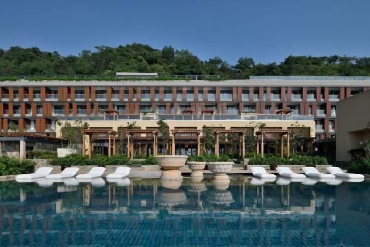 A hotel with a swimming pool, multiple balconies, and a forested hill in the background.