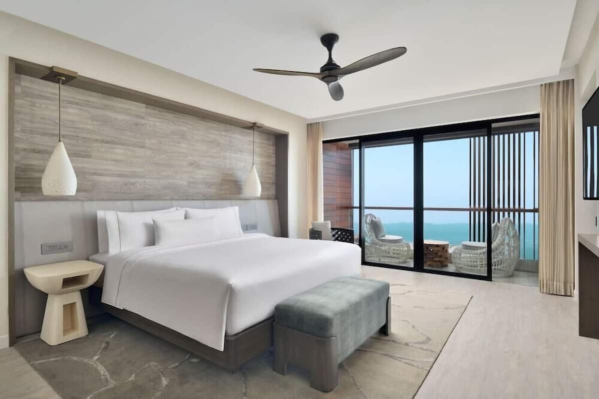 A modern hotel room with a large bed, a ceiling fan, a view of the ocean, and a balcony with wicker furniture.