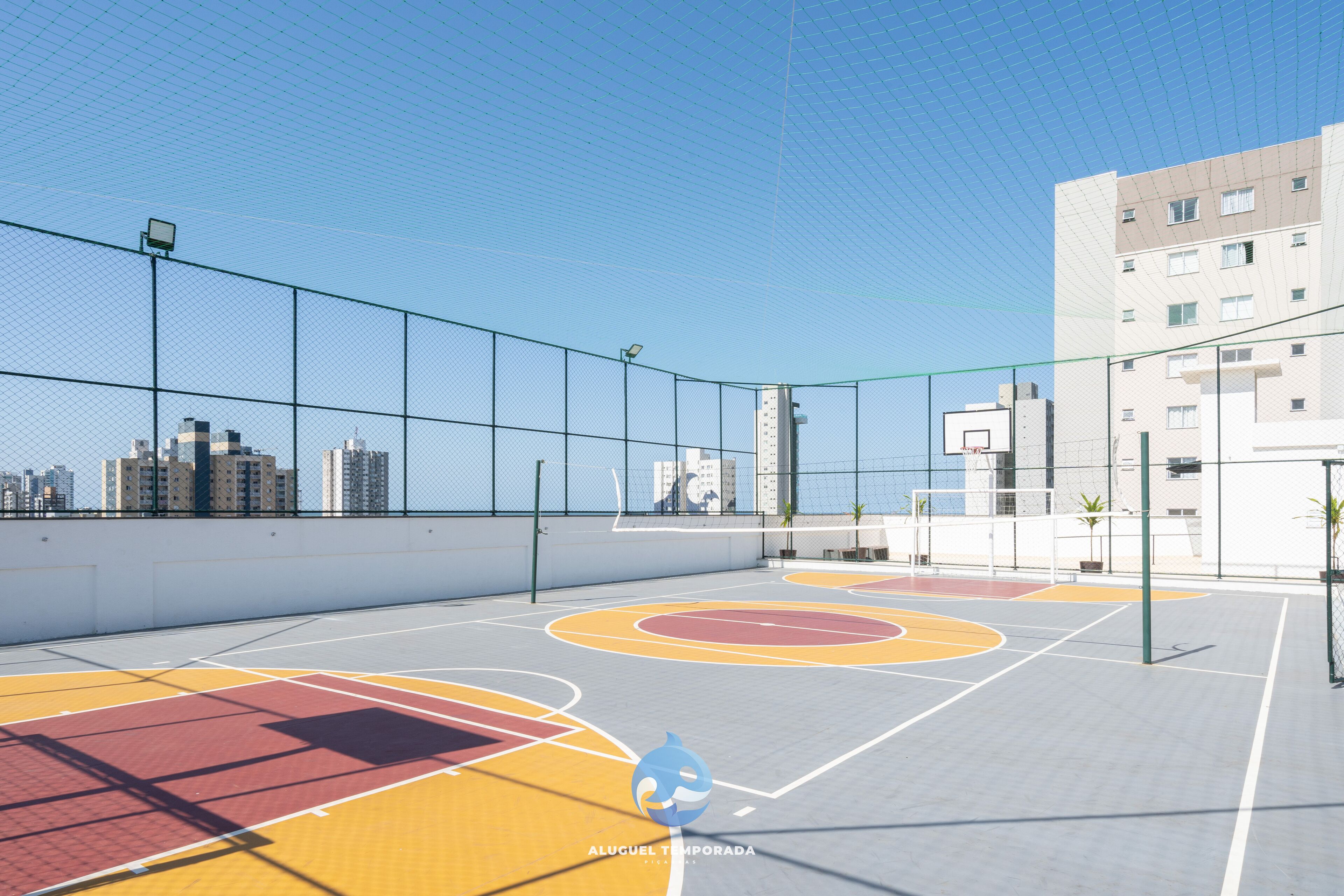 An outdoor basketball court with a net, surrounded by a fence, and buildings in the background.