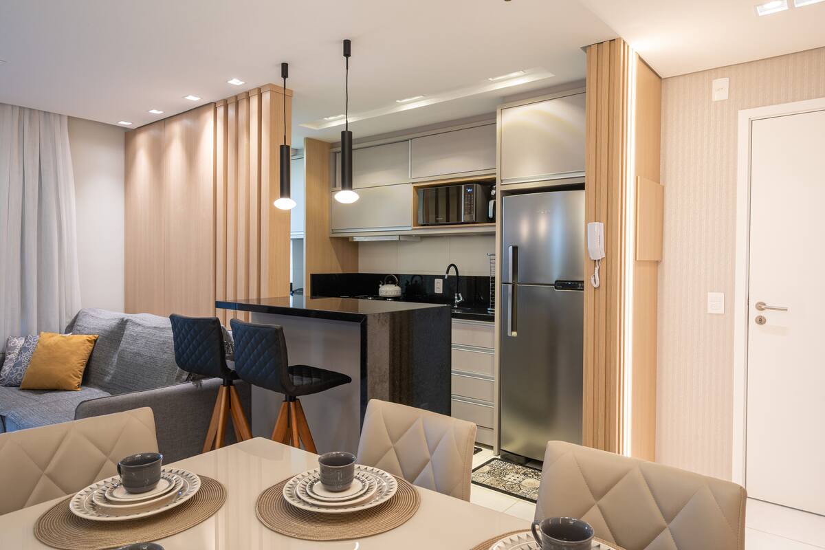 A modern kitchen with a refrigerator, microwave, and kettle, a dining area with a table and chairs, and a living area with a sofa and cushions.