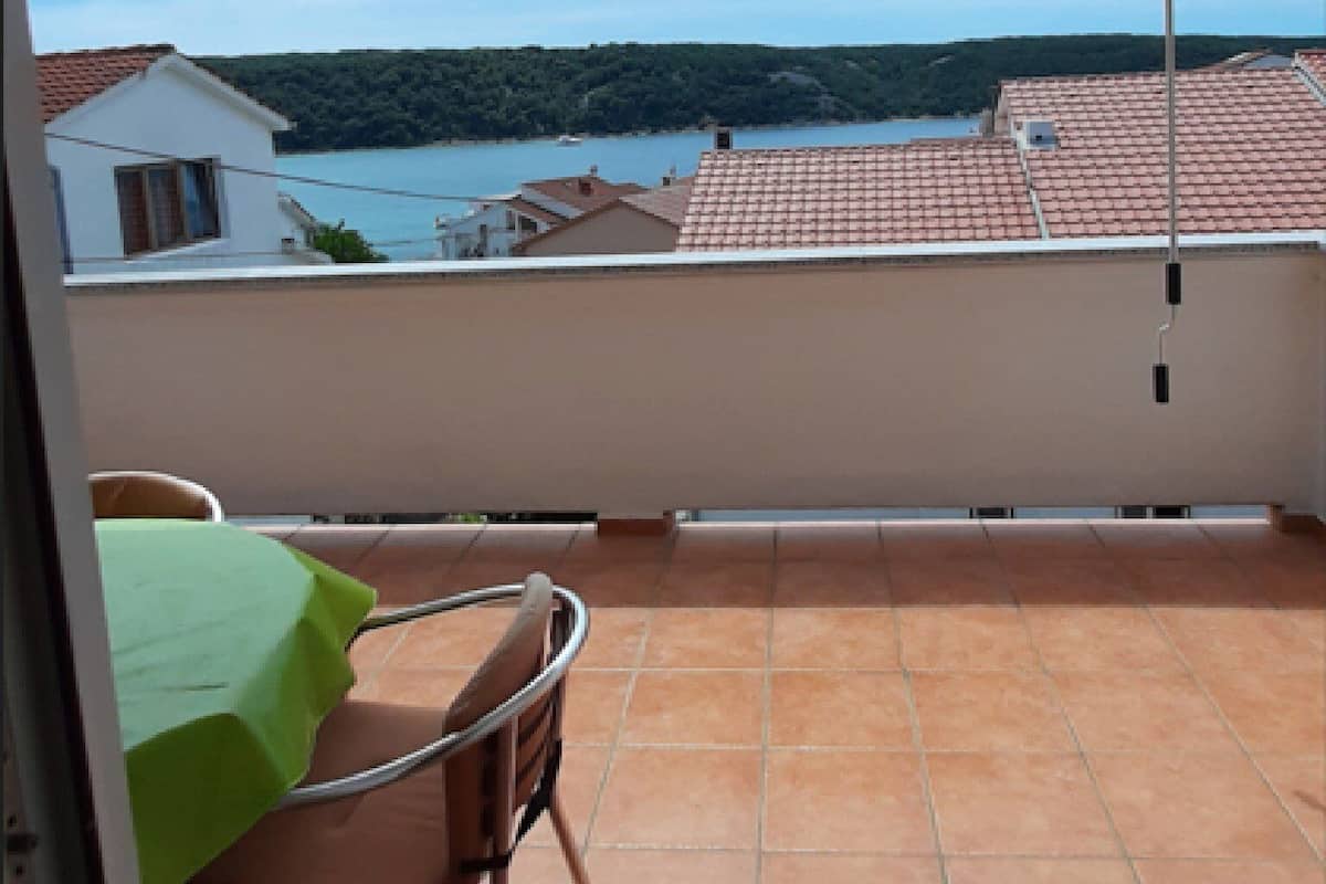 A balcony with a view of a lake and hills, a table with a green tablecloth, and a chair.