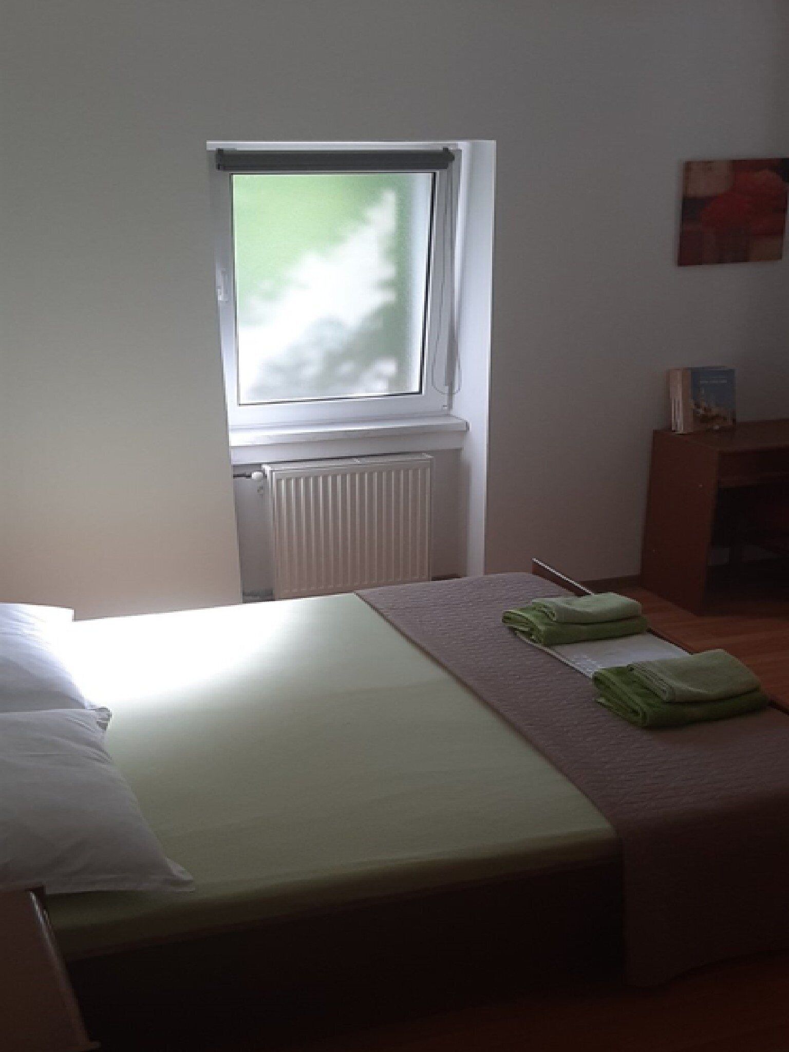 A bedroom with a single bed, a radiator, a window, and a small table with a book and a figurine.