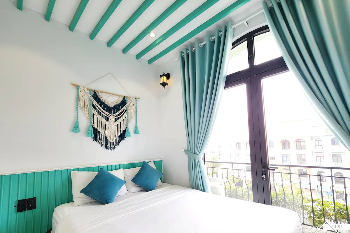 A hotel room with teal accents, a bed with white and blue bedding, a macrame wall hanging, and a balcony with a view of buildings.
