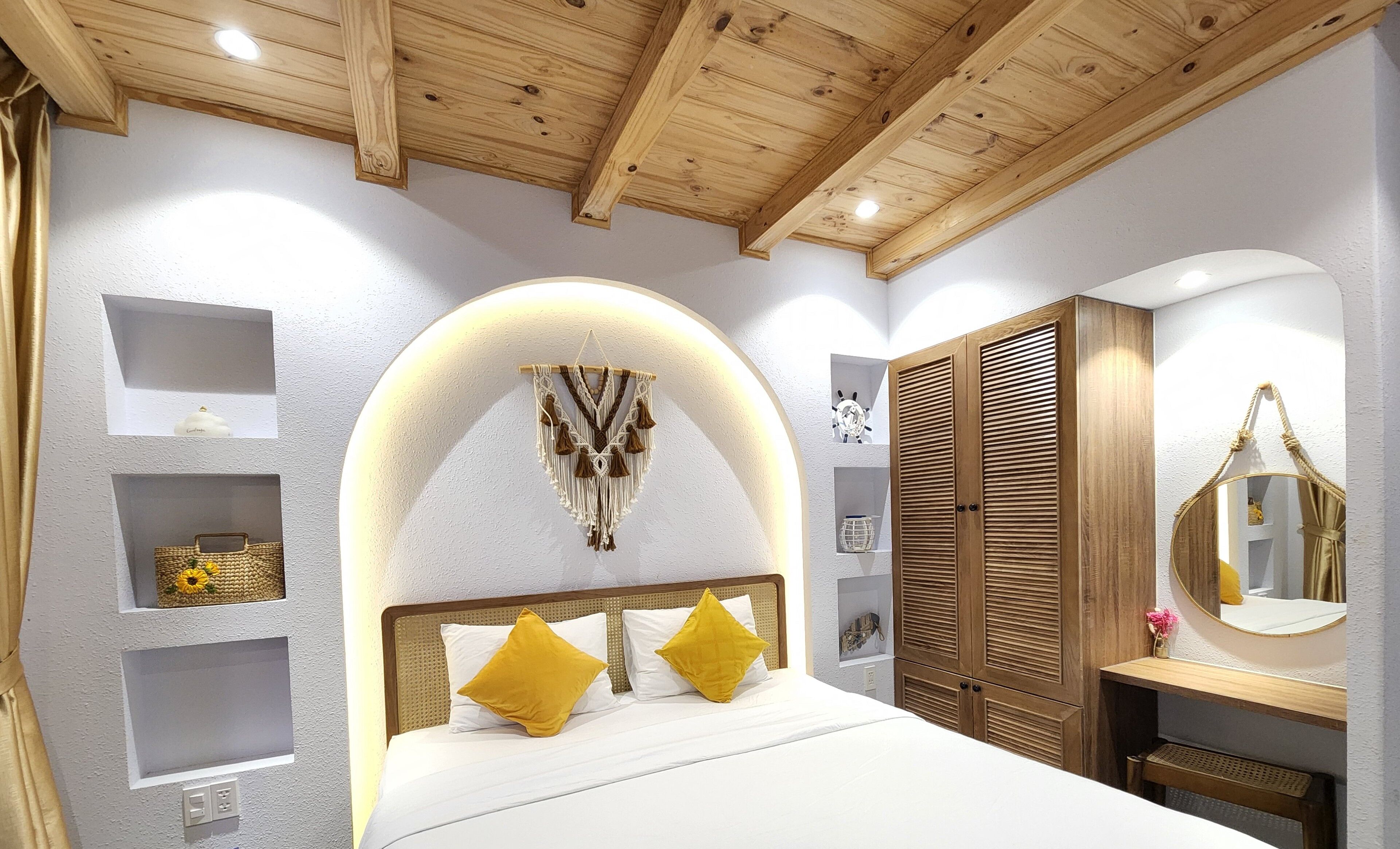 Een slaapkamer met een houten plafond, een bed met witte beddengoed en gele kussens, een houten garderobe en een spiegel met een touwgreep.