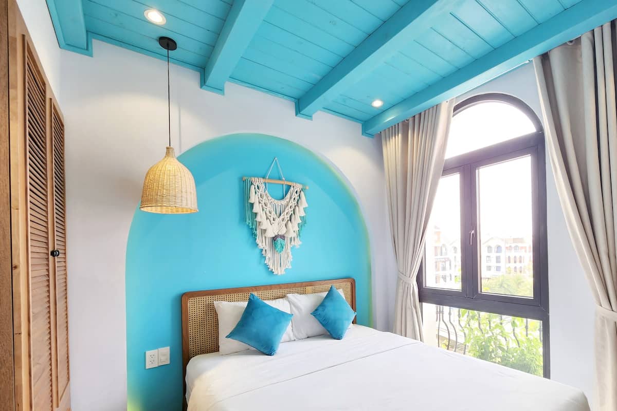 A bedroom with a bed, a macrame wall hanging, a wicker pendant light, and a window with curtains.