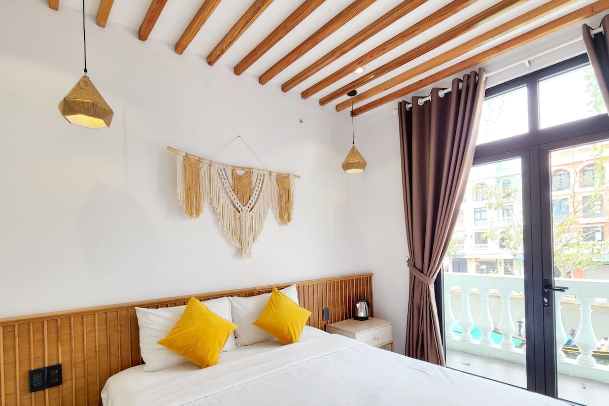 A hotel room with a wooden ceiling, a bed with white bedding and two yellow pillows, a macrame wall hanging, and a view of a balcony with a railing.