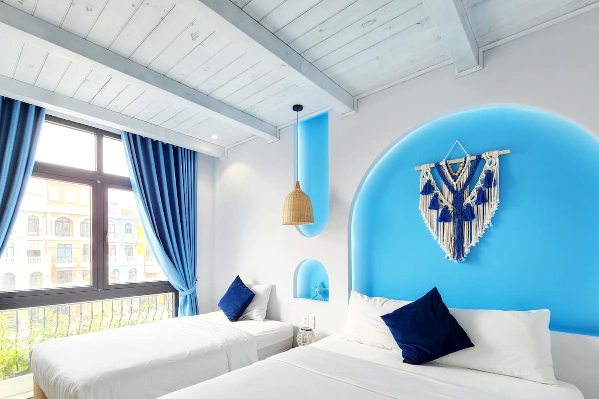 A modern bedroom with two beds, a large window with blue curtains, a blue accent wall, and a macrame wall hanging.