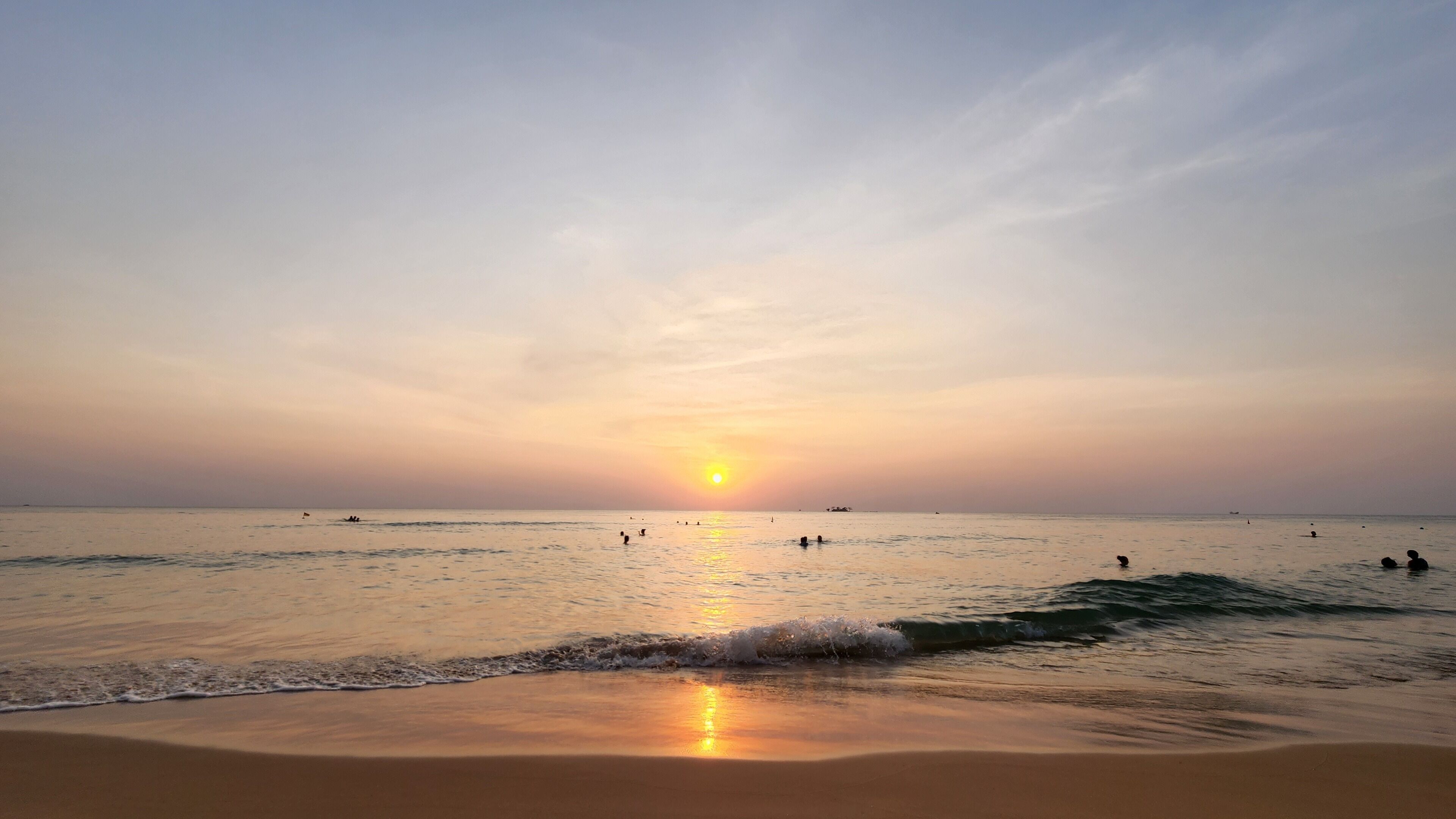 Una playa con el sol poniéndose en el horizonte, mar tranquilo y gente nadando.