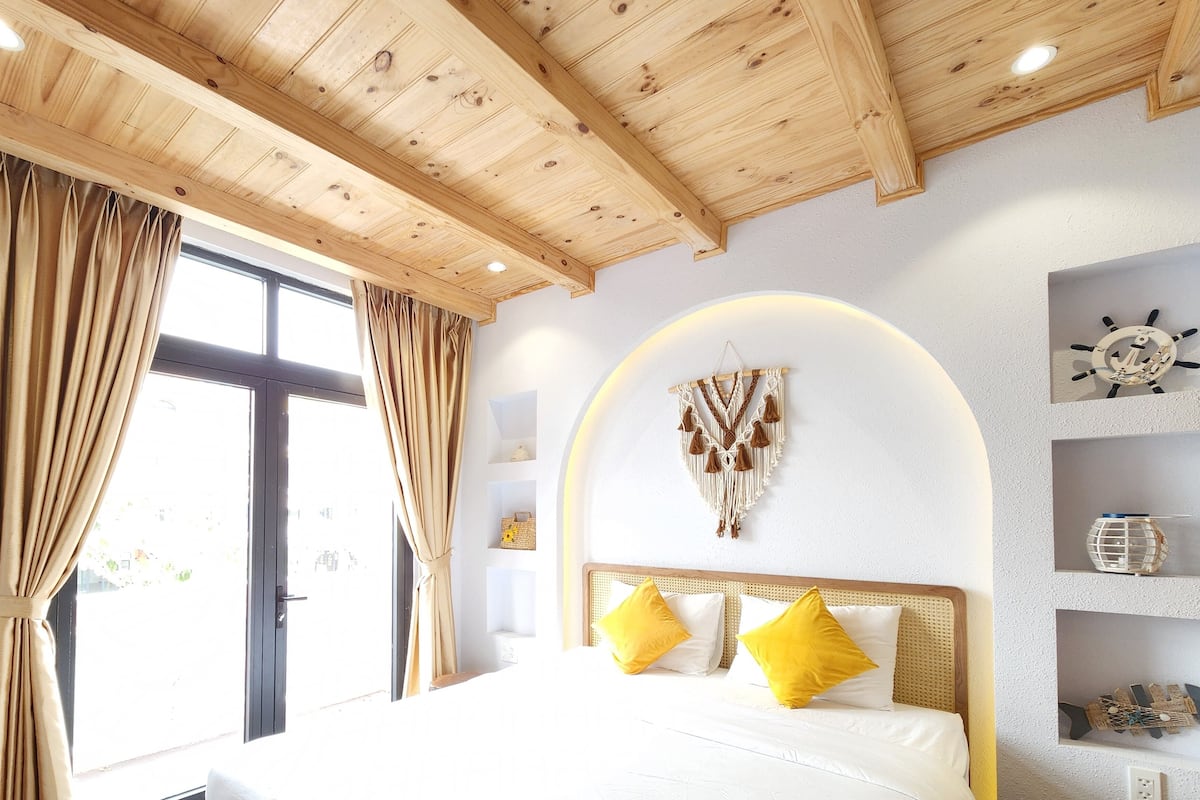 A bedroom with a wooden ceiling, a bed with white and yellow pillows, a wall-mounted macrame piece, and a ship's wheel decoration.