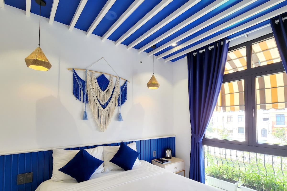 A hotel room with a bed, a nightstand, a wall hanging, and a window with curtains.