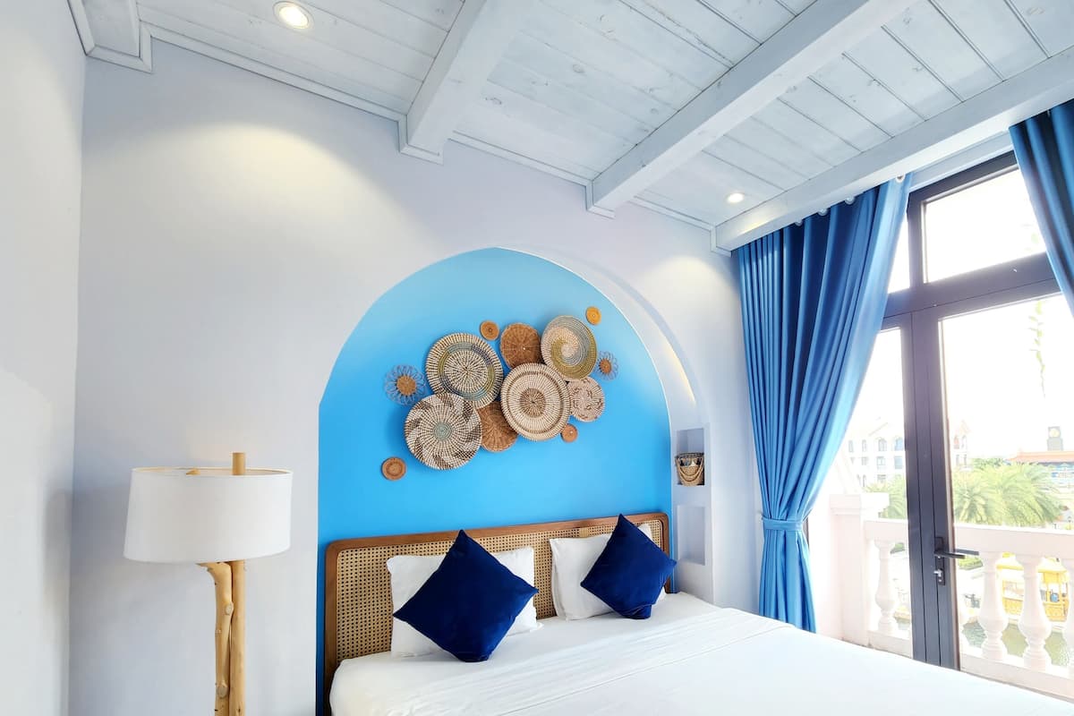 A hotel room with a bed, a wall-mounted lamp, a decorative wall piece, and a view of the outside through a window with blue curtains.