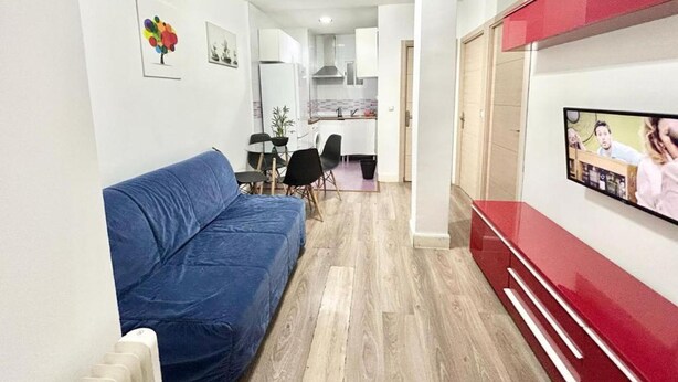 Un salón con un sofá azul, un televisor montado en la pared y una cocina al fondo.