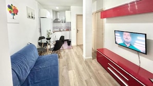 Una sala de estar moderna con un sofá, una mesa de comedor con sillas, una zona de cocina y un televisor montado en la pared.