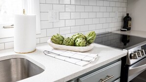 A kitchen with white subway tiles, a stainless steel sink, a bowl of artichokes, a roll of paper towels, and a black bottle on the counter.