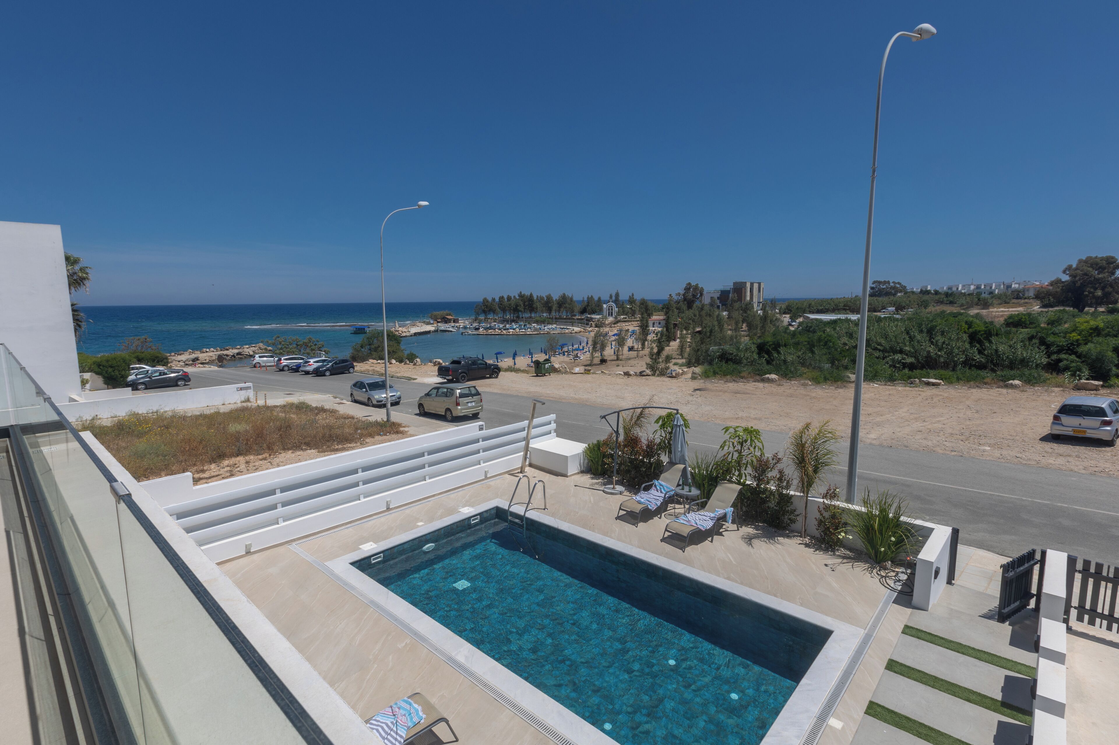 A swimming pool with a view of the sea, a road with parked cars, and a clear sky.