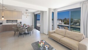 A modern living room with a sofa, dining table, and chairs. There is a kitchen area with cabinets and a countertop. Large windows offer a view of the outdoors. - Protaras Holiday Villa Ti39 (Paralimni)