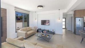 A modern living room with a sofa set, a coffee table with items, a flat-screen TV, and a kitchen area with a refrigerator. - Protaras Holiday Villa Ti39 (Paralimni)