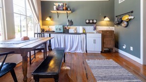Een keuken met houten vloeren, een eettafel en een bankje. Er is een raam met gordijnen, een wandplank met decoratieve objecten en een wandbevestigde panhanger.