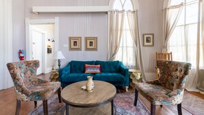 Een kamer met een blauwgroene bank, twee gestreepte fauteuils, een houten salontafel en een groot raam met gordijnen.