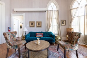 Una habitación con un sofá azul verdoso, dos sillones estampados, una mesa de centro de madera y un ventanal con cortinas.