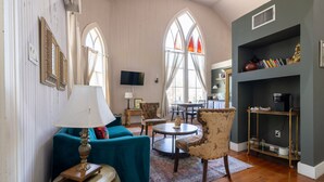 Een ruime woonkamer met een blauwgroene bank, een ronde salontafel en gestreepte fauteuils. Er is een boekenkast met decoratieve objecten en een flatscreen televisie aan de muur bevestigd.