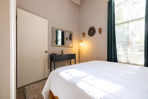 Ein Schlafzimmer mit einem Bett, einem Schminktisch, einem Spiegel, einer Wandleuchte und einem Fenster mit Vorhängen.