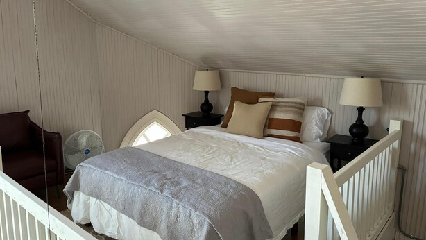 Een slaapkamer met een bed, twee nachtkastjes met lampen, een stoel, een ventilator en een raam.