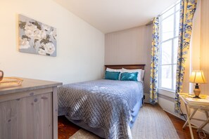 Un dormitorio con cama, mesita de noche, lámpara, ventana con cortinas y un cuadro en la pared.