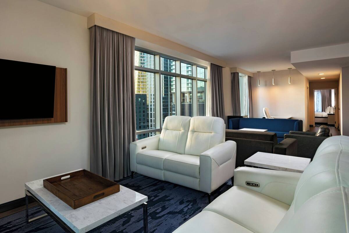 A modern hotel room with a large window, a sofa, a small table, and a TV mounted on the wall.
