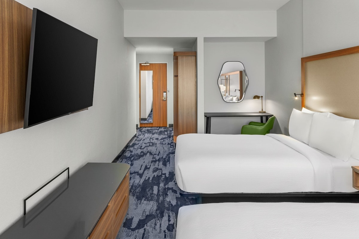 A hotel room with a flat-screen TV mounted on the wall, a bed with white bedding, a desk with a mirror, and a green chair.