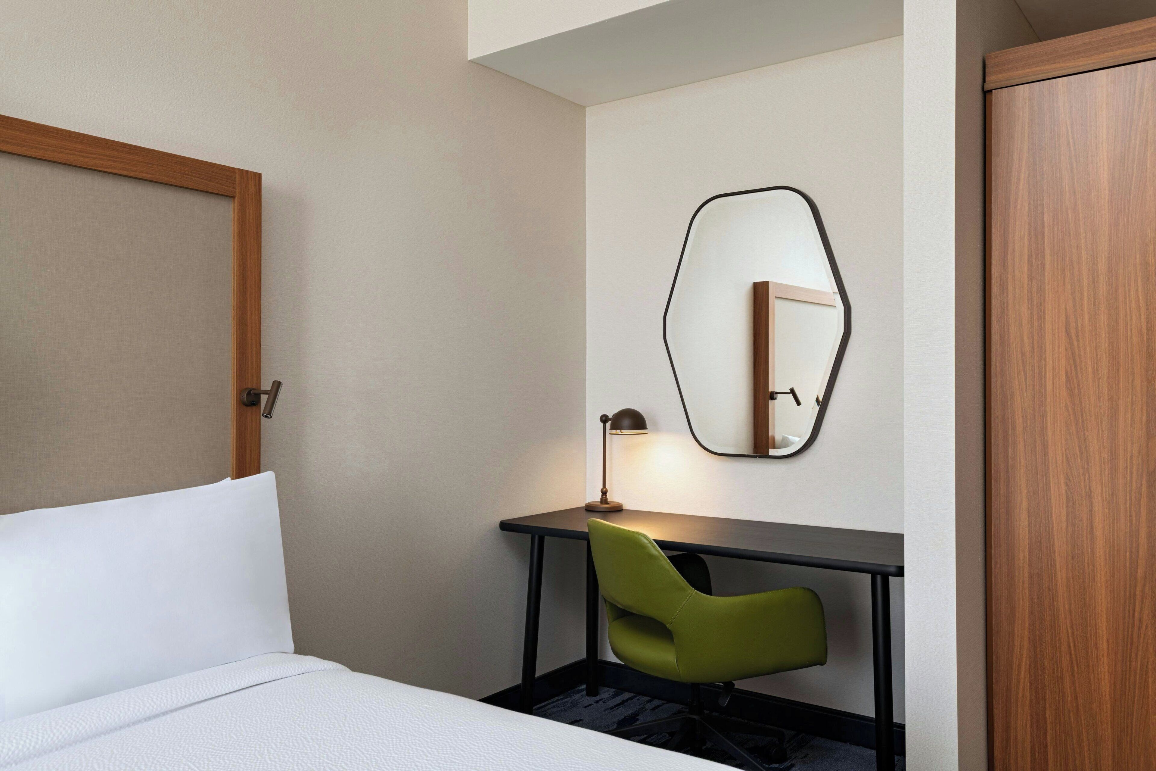 Een hotelkamer met een bed, een bureau, een groene stoel, een lamp en een spiegel aan de muur.