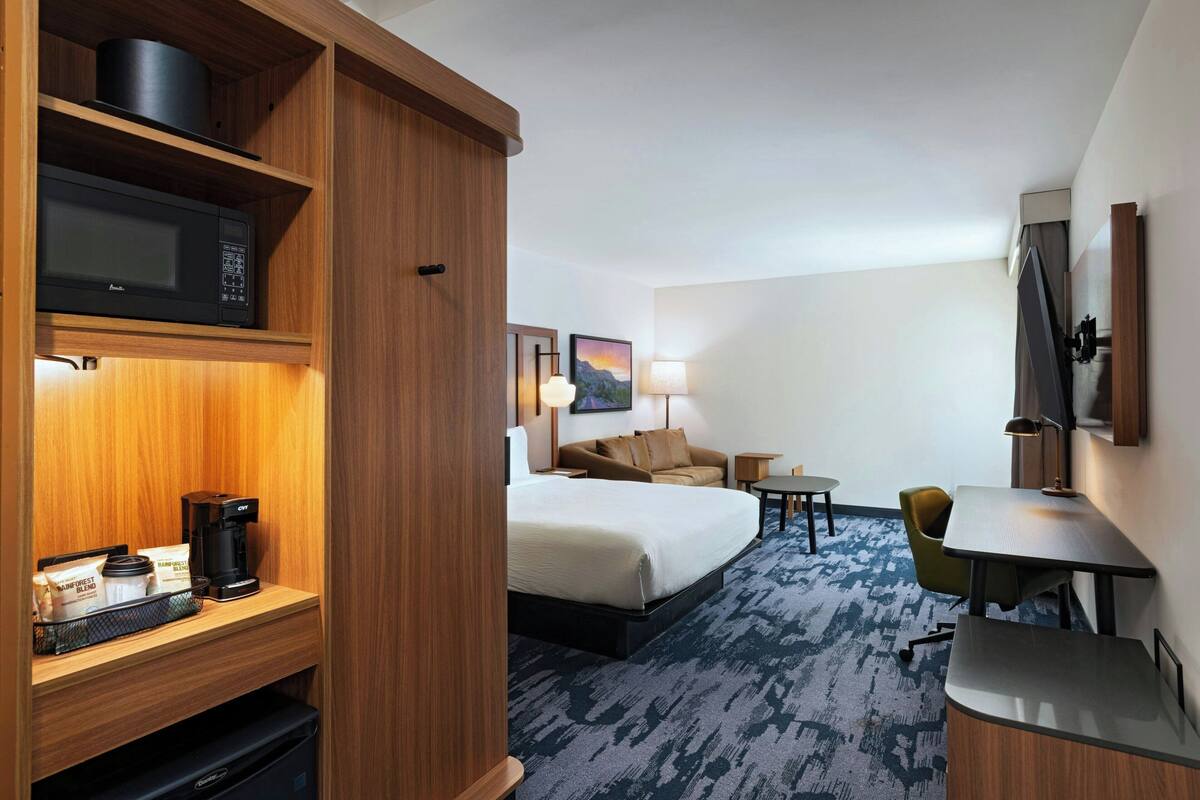 A hotel room with a bed, a desk, a chair, a microwave, a coffee maker, and a TV.