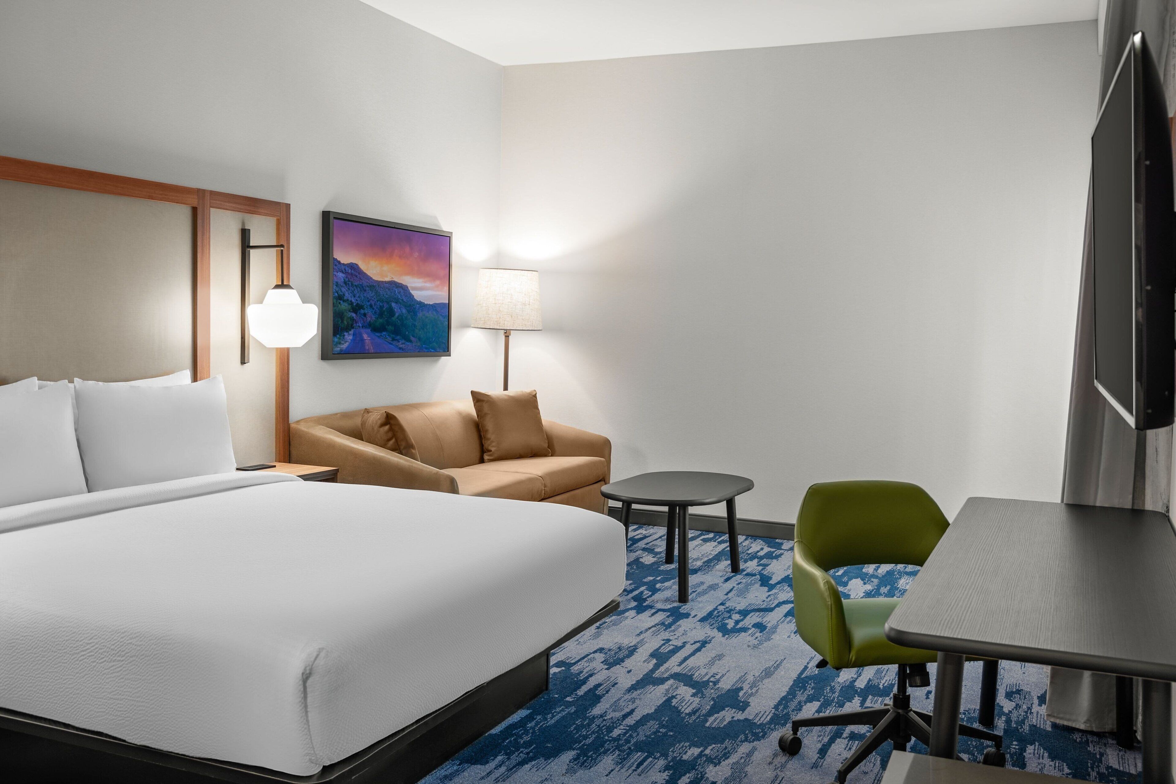 A hotel room with a large bed, a sofa, a small table, a green chair, a TV, and a painting on the wall.