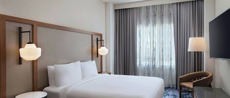 Habitación de hotel con una cama grande, dos lámparas de noche, un televisor, una silla y una ventana con cortinas.