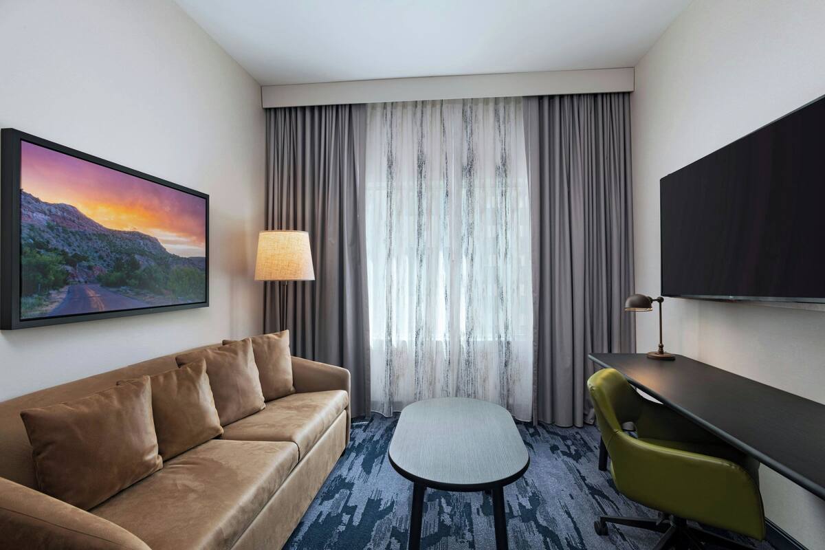 A hotel room with a sofa, a small table, a chair, a TV, and a window with curtains.